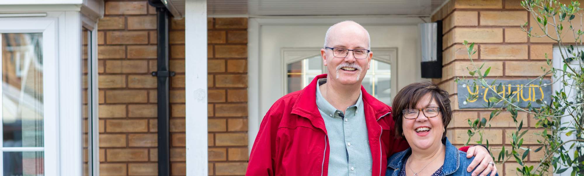 A man and woman standing together with arms around each other outside their home